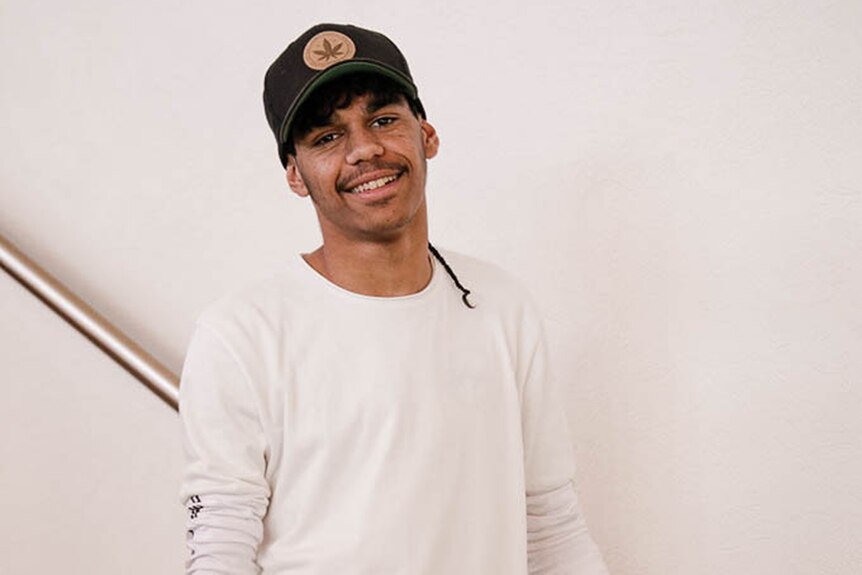 Colour photo of Kyle Lynch standing in white long-sleeve shirt in front of white wall holding onto steel staircase banister.