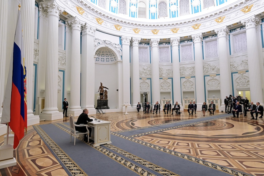 Putin sits at a desk in a huge domed room with people on chairs at the other side facing him