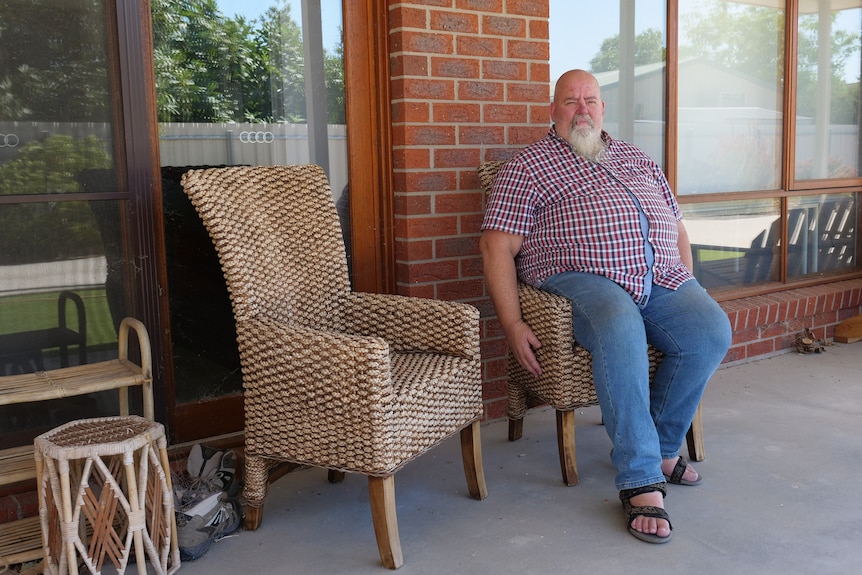 A bald man with gray beard and red and blue plaid shirt and blue jeans sits in a cane chair