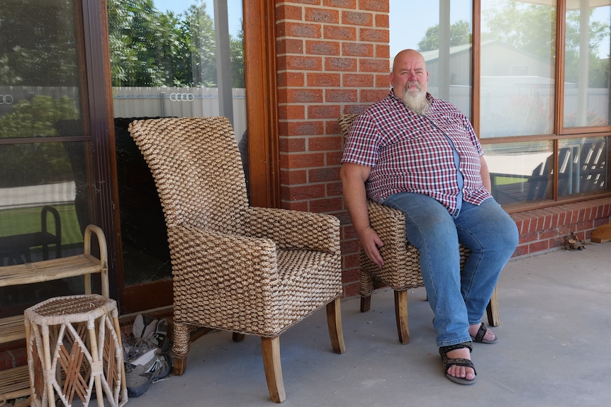 Un homme chauve avec une barbe grise et une chemise à carreaux rouge et bleu et un jean bleu est assis dans une chaise en rotin