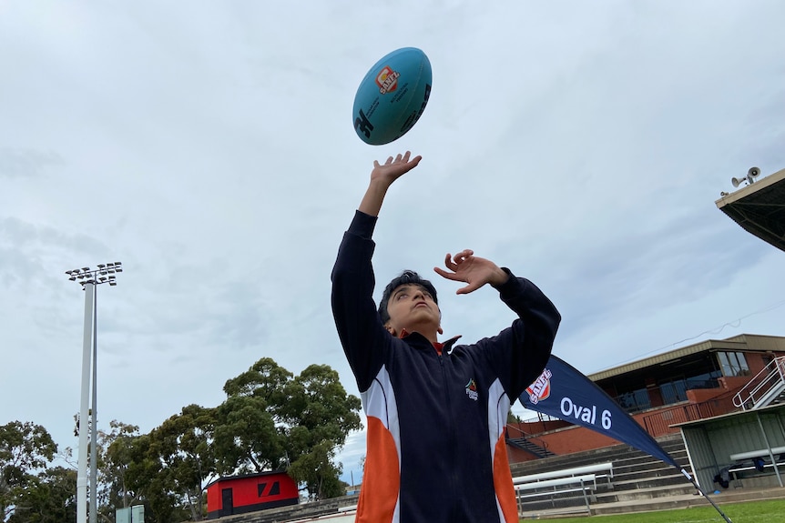 A teenage boy in a dark jacket tossing a football in a footy oval