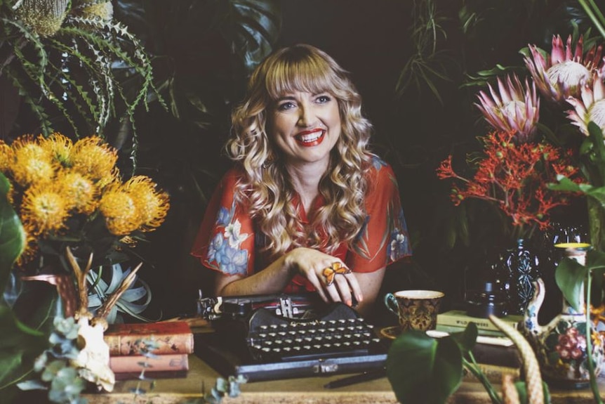Colour photo of writer Holly Ringland sitting as a desk with a type writer and surrounded by flowers.