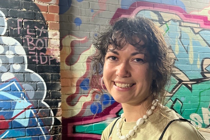 A young woman with dark curly bangs smiling in front of a colourful spray painted wall in an outdoor dining area. 