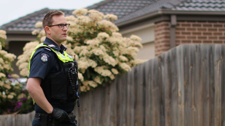A male police officer stands in the front yard of a suburban home, in front of a wooden fence and garden bed.