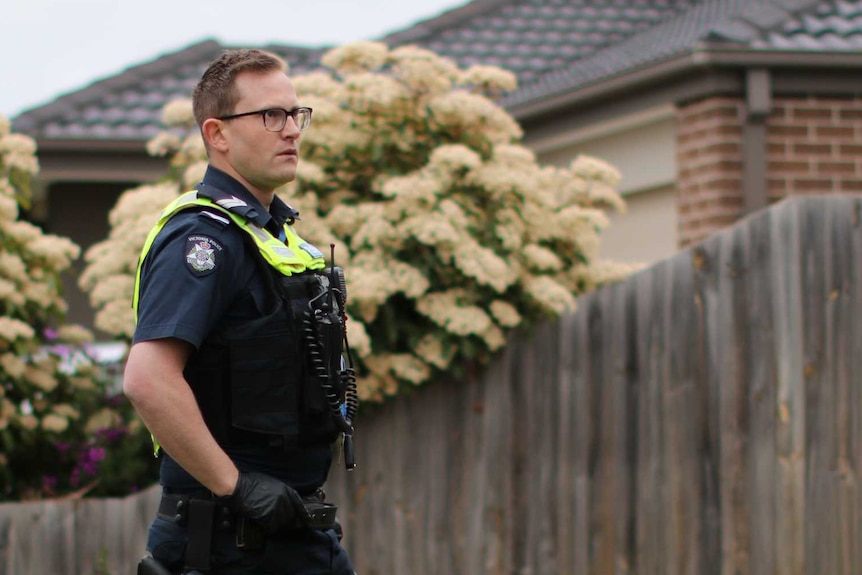 A male police officer stands in the front yard of a suburban home, in front of a wooden fence and garden bed.