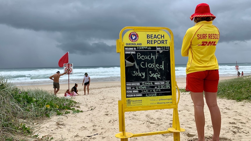 a life saver in uniform looks from the beach out at the surf with dark clouds above next to a sign saying the beach is closed
