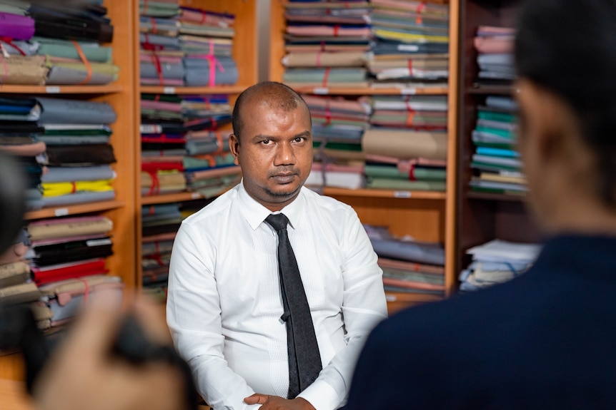 A man looks at the camera seated in front of bookshelves.