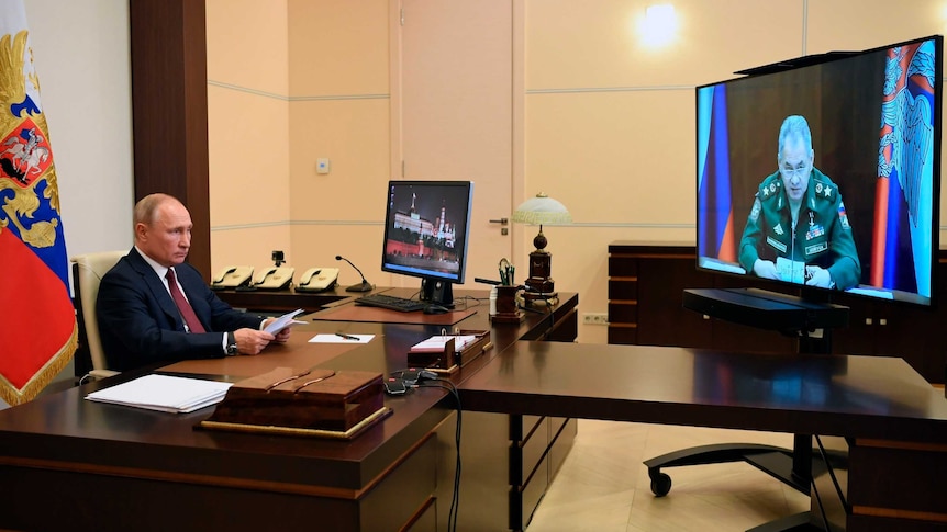 Russian President Vladimir Putin sits at his desk looking at TV screen that shows a video conference with the Defence Minister.