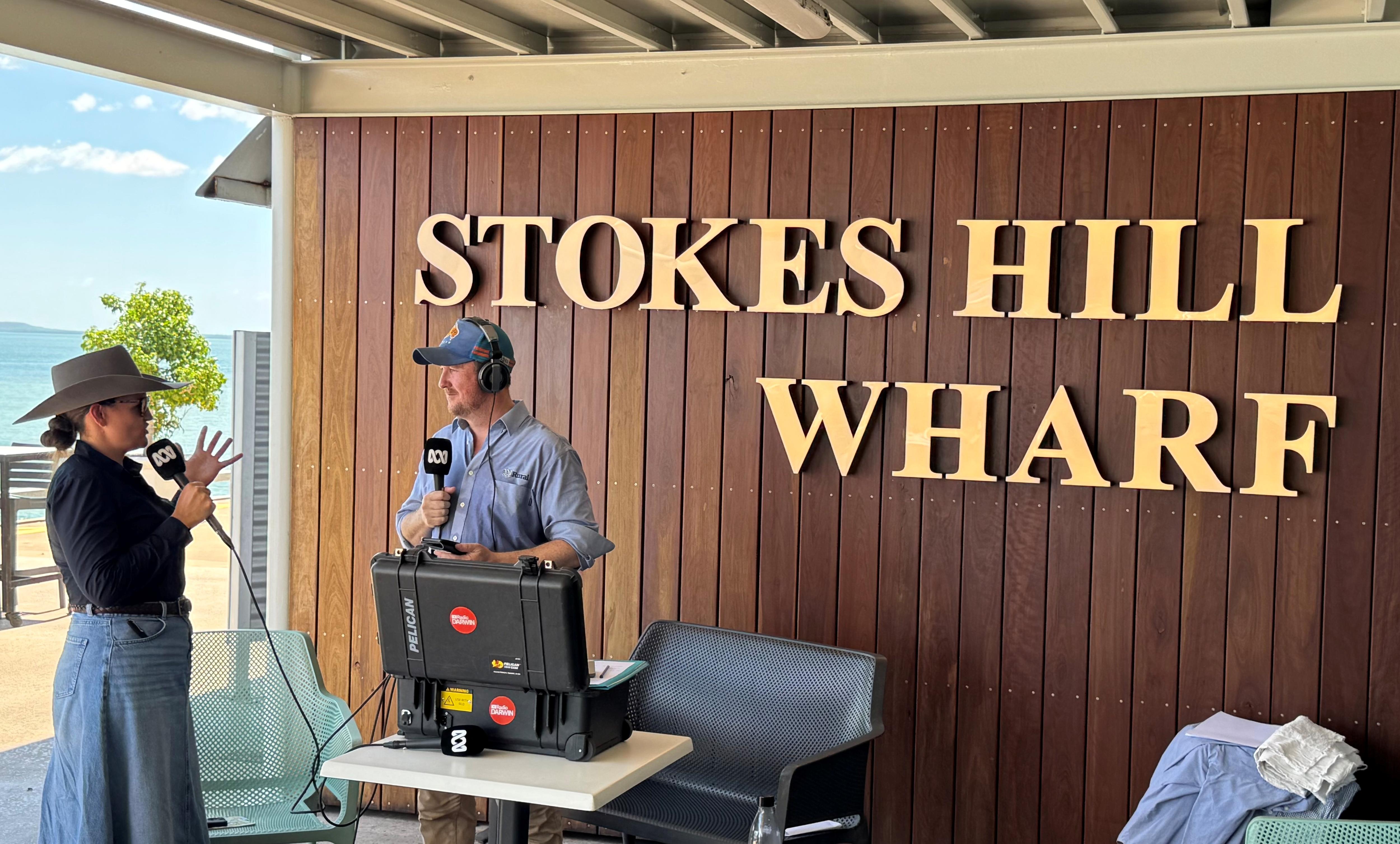 a woman with bush hat holding a microphone speaks with a man with a radio microphone in an veranda setting