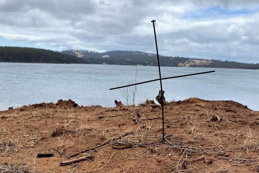 Photograph of a metal cross on a dry headland with more land visible on across a calm bay