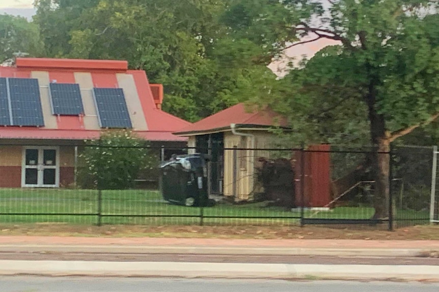 A wide shot of a school yard with a small black car sitting on its side on the grass in front of buildings.