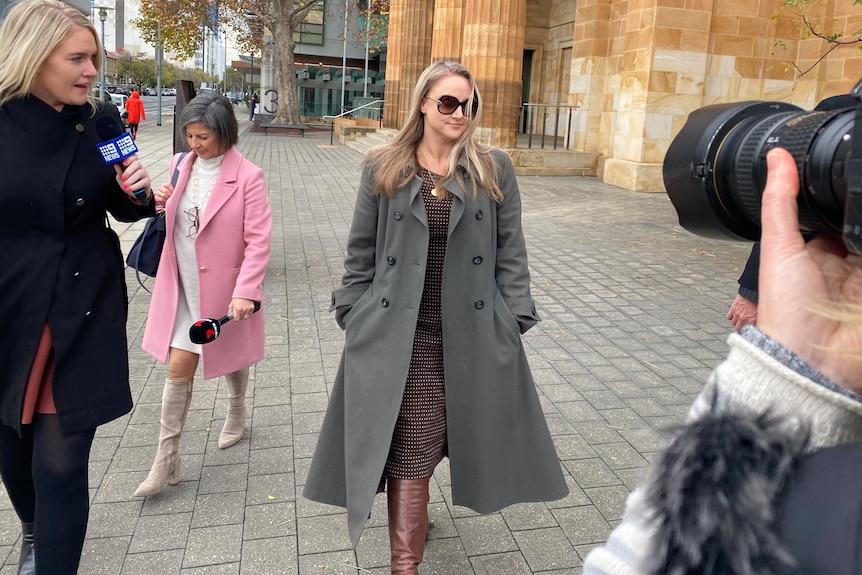 A woman wearing a long grey coat walks past reporters and a camera operator