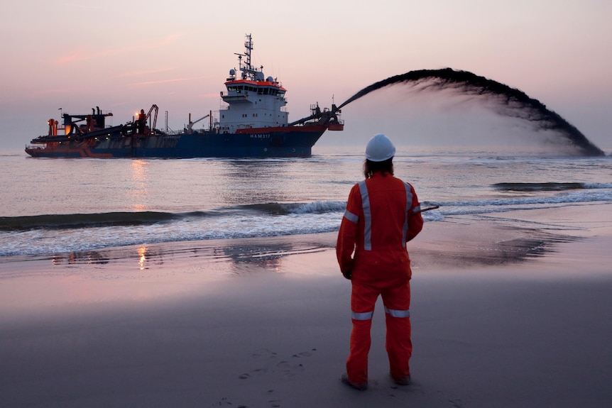 A person watches from the shore as a ship pumps sand onto the beach.