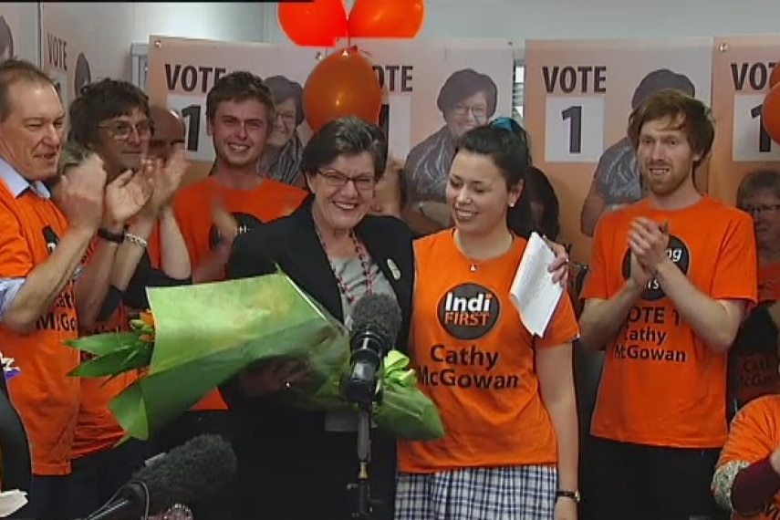 Cathy McGowan holds a bunch of flowers while campaign workers in orange t-shirts stand with her and applaud.