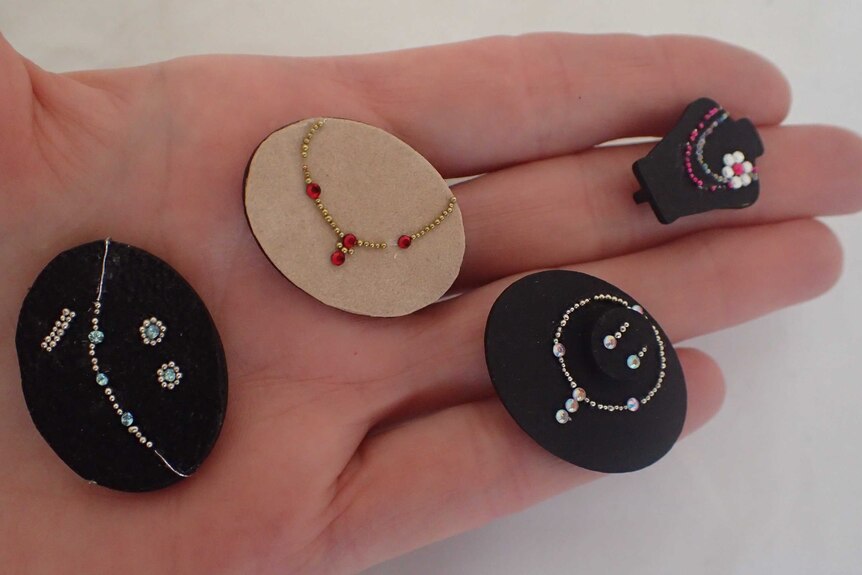 A hand holds four miniature jewellery pads displaying miniature necklaces.