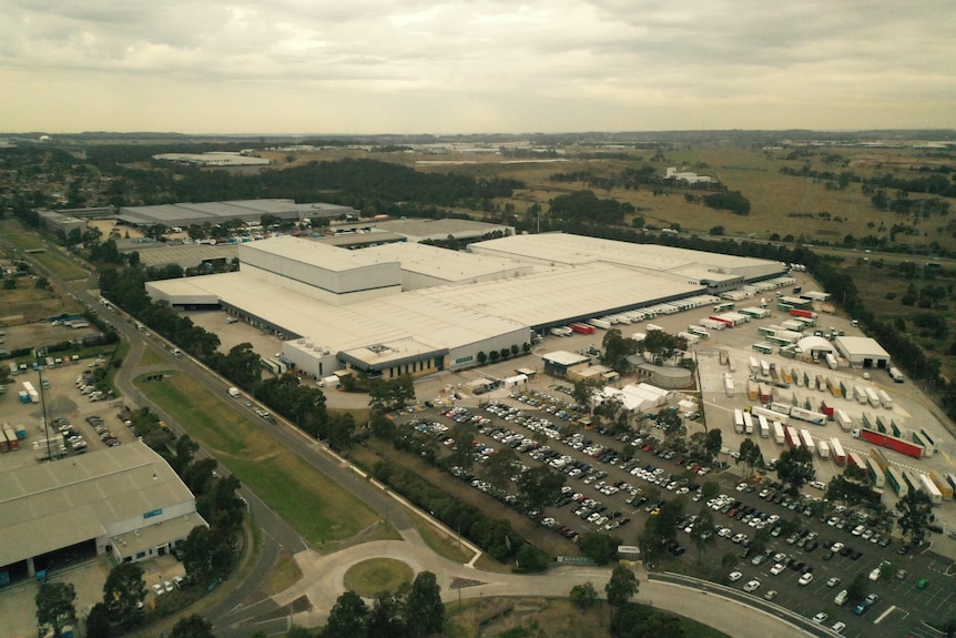 An image taken from a drone of a large industrial building in a suburban area with a car park filled with trucks and cars.