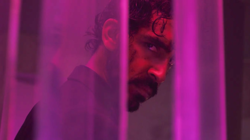 A film still of Dev Patel close-up. He has an angry expression, and has facial wounds, and can be seen looking through a gap.
