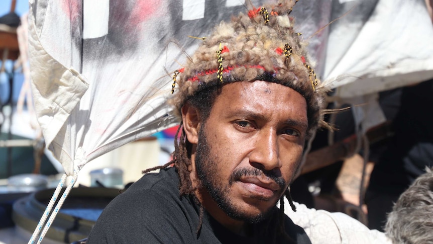 Papua New Guinean man wearing traditional hat in front of refugee flag.