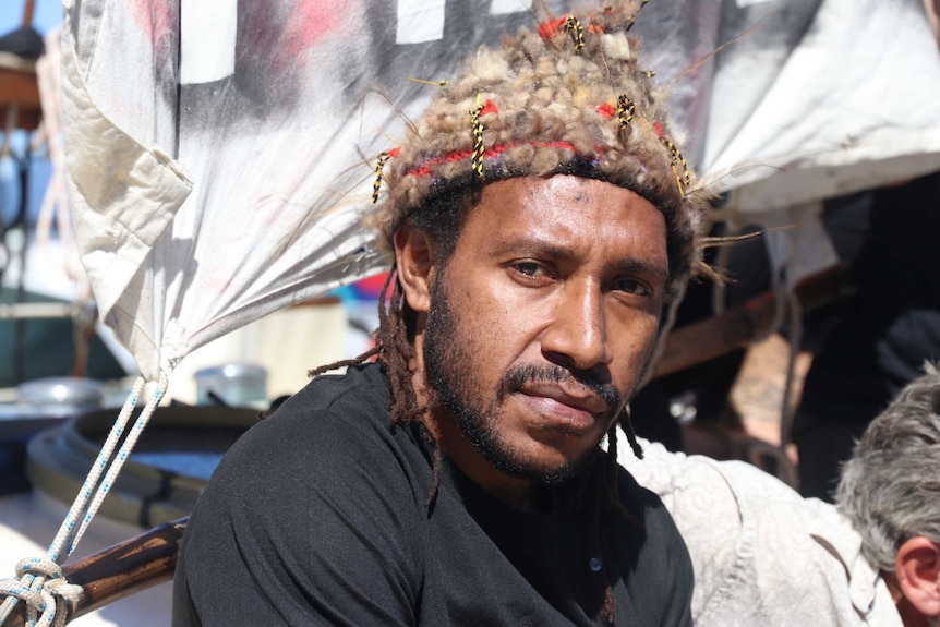 Papua New Guinean man wearing traditional hat in front of refugee flag.