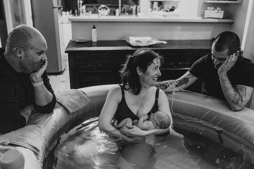 A woman with a new born baby sits in a blow-up pool with two man on either side.