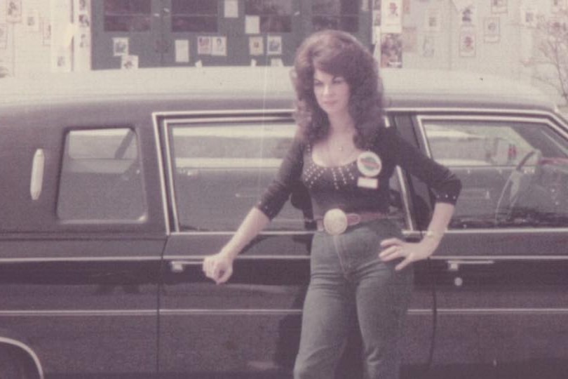 a vintage photo of a woman with big hair and big gold belt buckle leaning on a black car in the 1980s