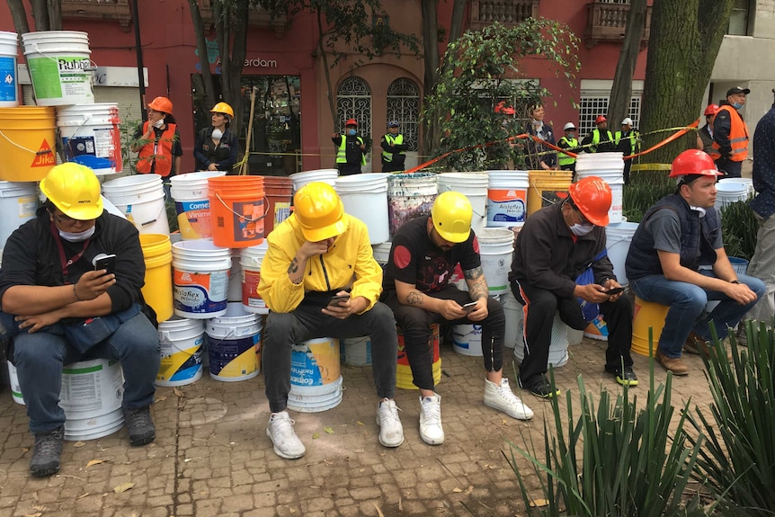 Relatives and neighbours, wearing hard hats, sit on buckets as they watch rescue workers.