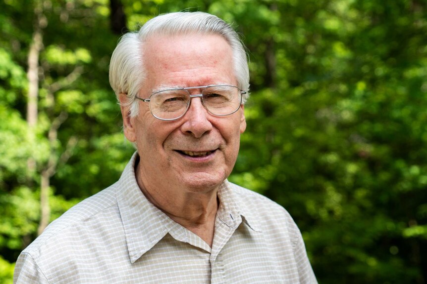 An older man in glasses stands in front of some trees