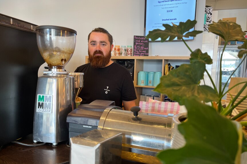 A bearded man stands behind a coffee machine in a cafe.
