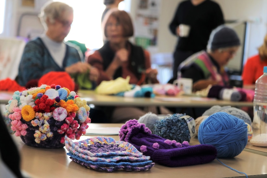 Brightly coloured beanies, yarn, crocheted coral and crocheted squares are spread across a table, with ladies knitting beyond.