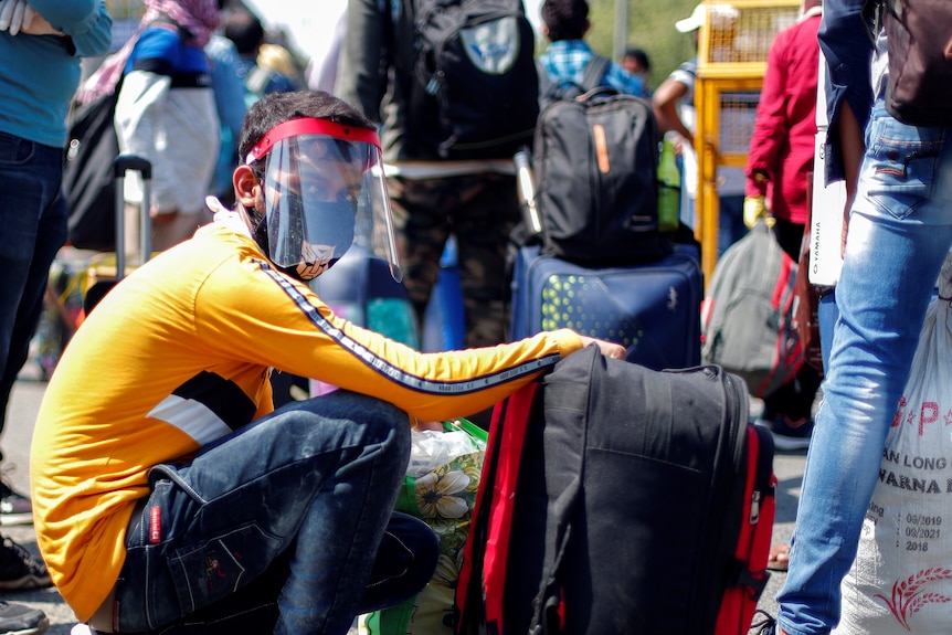 A man wearing a face shield  squats next to his belongings among a crowd of people