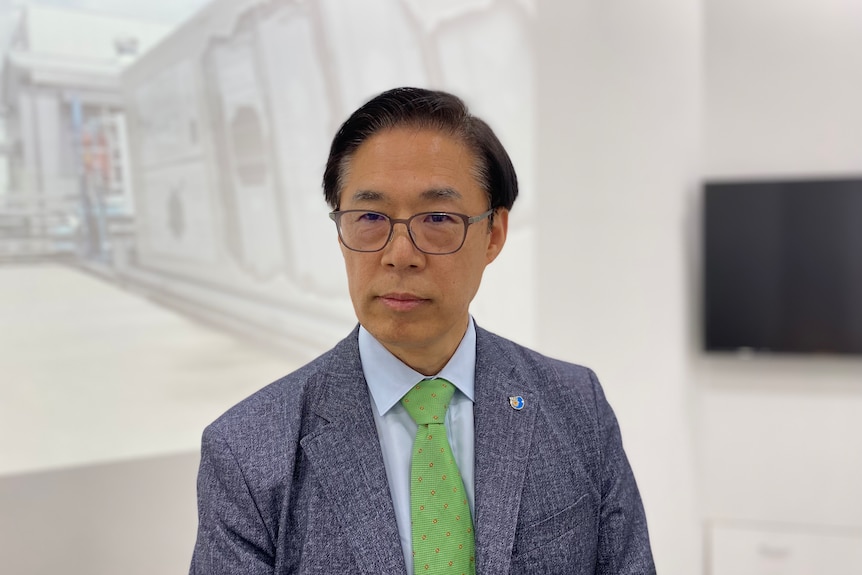 An older Korean man wearing a suit and tie and glasses stands in the foyer of a building.