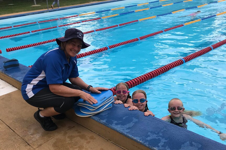 A woman kneels by the pool alongside kickboards with children in the pool and they smile at the camera