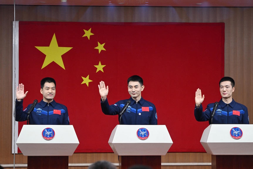 Three male Chinese astronauts wave as they stand in uniforms before a huge Chinese flag.
