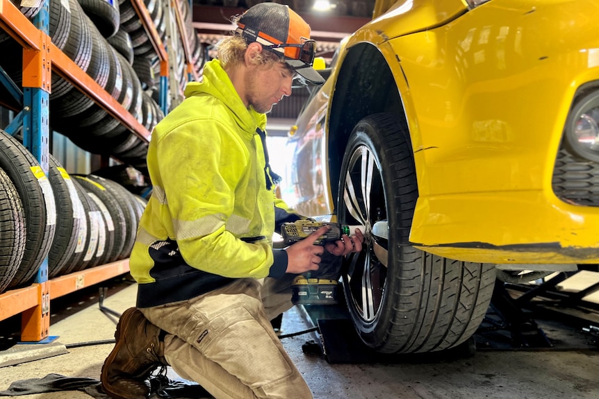 A man wearing high-vis kneels next to a yellow Mustang changing its tyres.