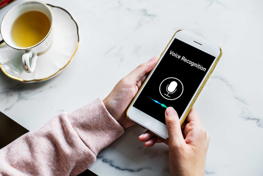 Woman holds a phone on a white marble table that says 'voice recognition' on the screen.