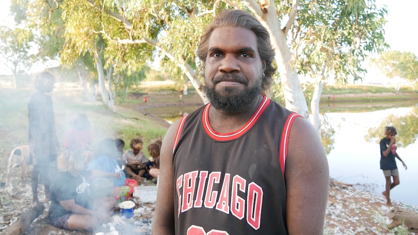 Felix Williams faces the camera in front of a background of kids eating bush tucker near a fire at the Balgo dam.