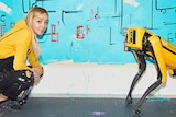 A middle-aged woman with long blonde hair crouches in front of a large blue painting, smiling. Near her is a yellow robot dog.