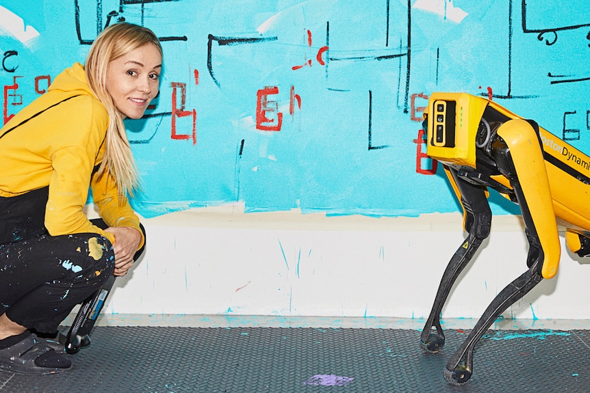 A middle-aged woman with long blonde hair crouches in front of a large blue painting, smiling. Near her is a yellow robot dog.
