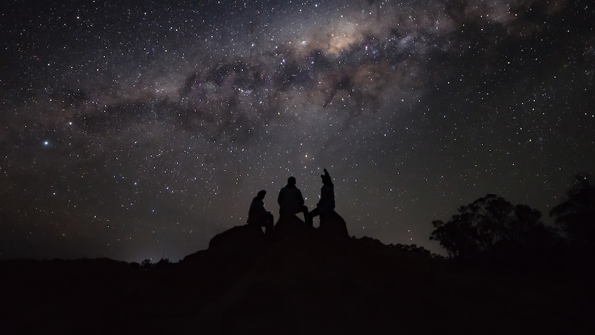 Nighttime photo of 3 people looking up at the milky way. They are silhouetted by the light from the stars.
