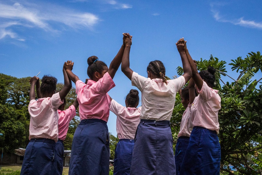 A small group of schoolgirls wearing pink shirts and navy skirts stand in a circle and raise their hands together, outside.