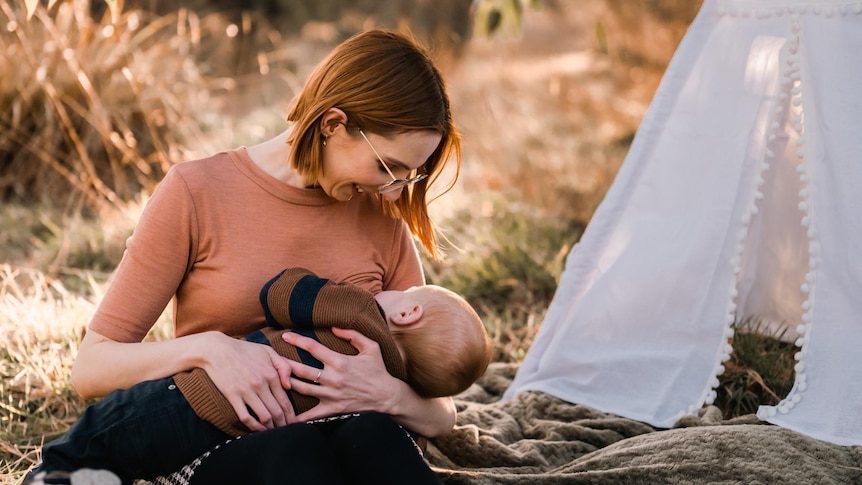Katya Daniel sits on the ground beside a white tent on a sunny day and breastfeeds her baby.