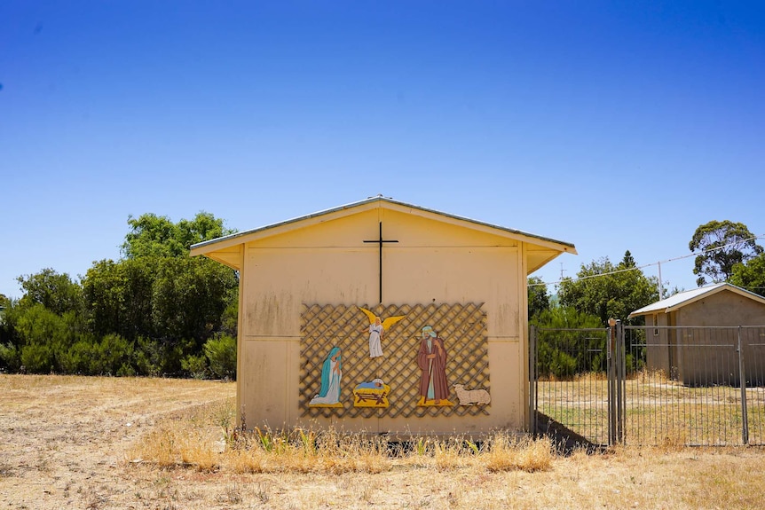 An old cream building on some unkept grass lawn with a cross and small nativity display on the wall.