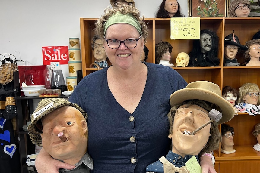 Woman holding two fake heads and standing in front of a cabinet full of heads