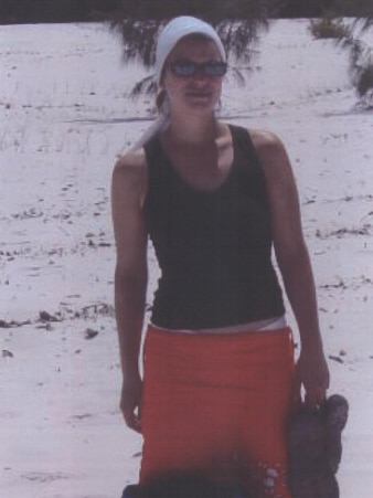 A woman in a black sleeveless shirt, red skirt and white headband on a beach with a backpack.