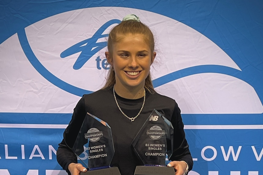 Young woman with a vision impairment stands smiling, holding two tennis trophies, blue and white tennis logo behind. Hair tied.