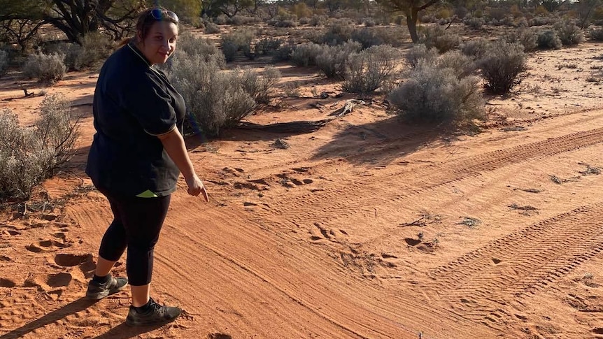 An Aboriginal woman wearing black leggings and shirt points to tracks in the brown dirt at a reserve.