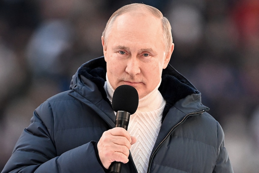 Vladimir Putin, wearing a thick navy puffer coat over a cream knitted rollneck jumper, holds a microphone to his mouth
