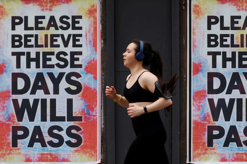 A woman wearing headphones runs past two signs saying "Please believe these days will pass".