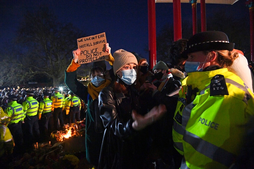 A woman wearing a face mask an beanie reacts as people gather at a protest.