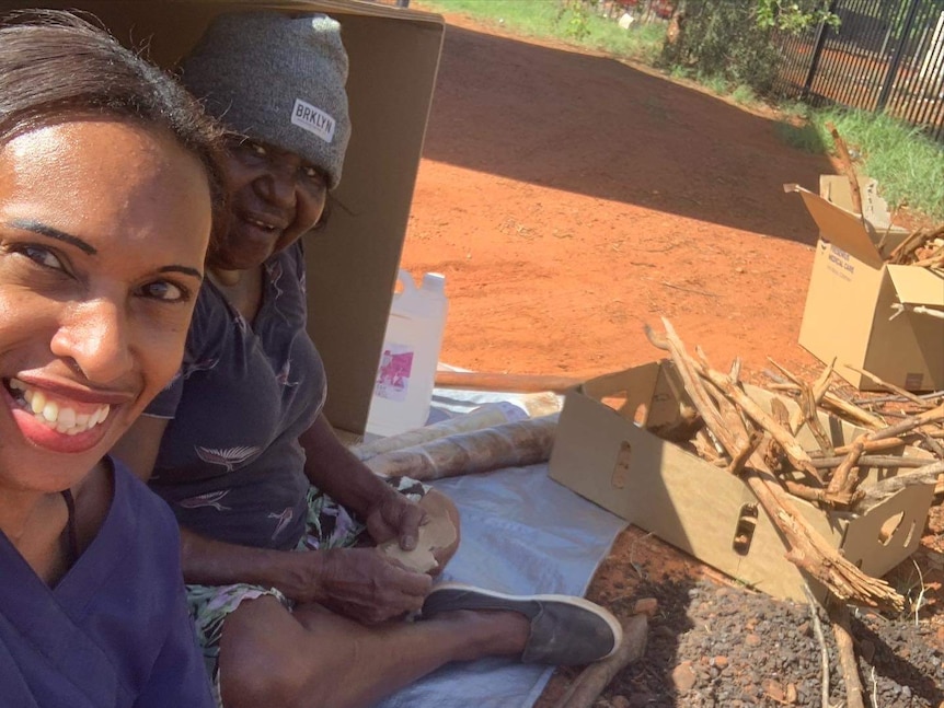 Two women smiling and sheltering under a box in front of a campfire surrounded by red dirt.